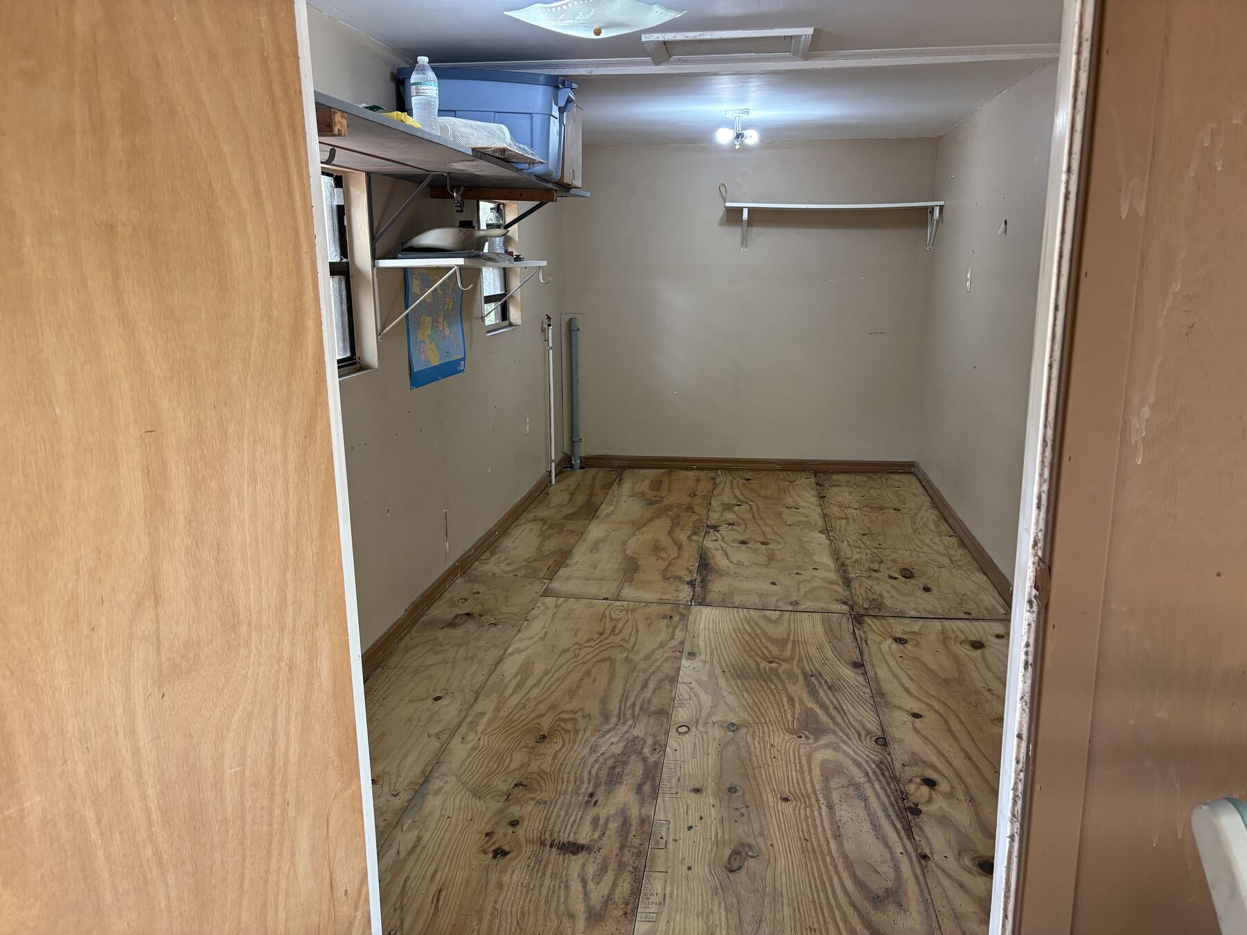 Looking in from the open door: bare plywood sheets laid over the old floor, two windows on the left wall, the washer standpipe in the corner
